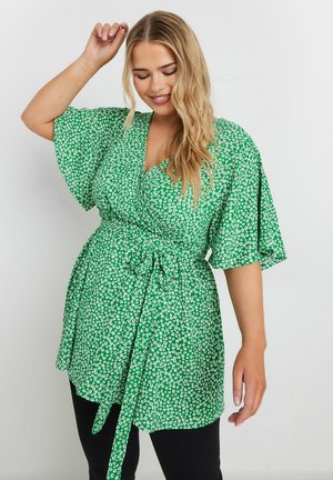 Blusa verde envolvente con patrón floral blanco, mangas cortas con vuelo, un cinturón para anudar y escote en V. Combinada con pantalones negros.