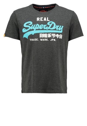 Mies, jolla on päällään tummanharmaa "SuperDry"-t-paita, mustat farkut, harmaat lenkkarit ja musta reppu, seisoo käsivarret taskuissa.