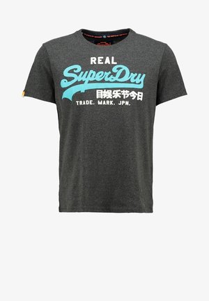 Mies, jolla on päällään tummanharmaa "SuperDry"-t-paita, mustat farkut, harmaat lenkkarit ja musta reppu, seisoo käsivarret taskuissa.