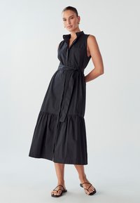 Abito in cotone nero con design a colletto, stile senza maniche, cintura in vita e orlo a strati. Presenta una chiusura frontale con bottoni e uno spacco laterale.