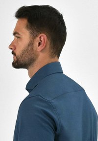 Blauwe overhemd met knoopsluiting en een gestructureerde kraag en gladde textuur, getoond op een man in zijprofiel, wat de pasvorm en het ontwerp van het shirt benadrukt.