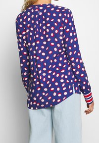 Topp med långa ärmar i blått med vita/röda ovala prickar. Har en veck bak och randiga manschetter för extra detalj.