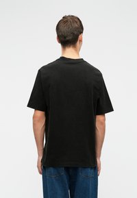 Muškarac s kratkom smeškom kestenjastom kosom nosi običnu crnu majicu kratkih rukava i plave traperice, stoji okrenut leđima na beloj pozadini.