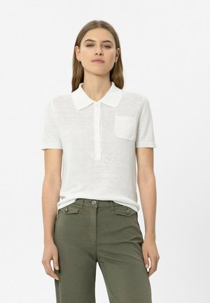 Woman wearing a white short-sleeve collared shirt with pocket and olive green high-waisted pants standing against a plain background.