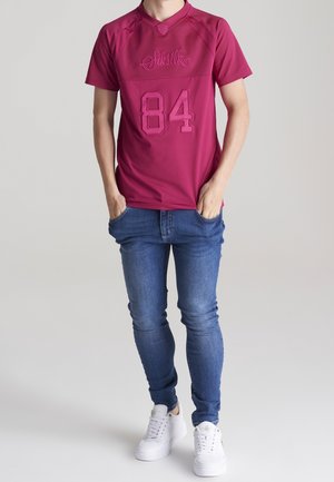 Camiseta deportiva de color granate con mangas cortas, cuello redondo y número "84" en relieve. Combinada con vaqueros azules y zapatillas blancas.