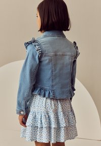 Ragazza vista di spalle con una giacca di denim chiaro con volant e una gonna bianca con motivo floreale blu e volant a strati.