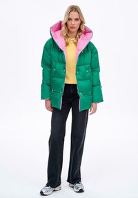 Zelená puffer bunda s růžovou podšívkou, patentními knoflíky a velkou kapucí. Nositá přes žlutý svetr a černé džíny, s volným střihem.