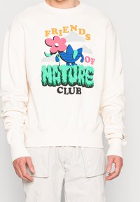 Persona vistiendo un sudadera crema con un gráfico colorido de un gato azul, una flor rosa, hierba verde y el texto "Amigos del Club de la Casa."