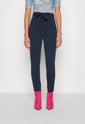 Kobieta w granatowych spodniach z wysokim stanem i wiązanym paskiem, w połączeniu z jasnoróżowymi botkami za kostkę oraz marszczonym, krótko rękawowym topem.