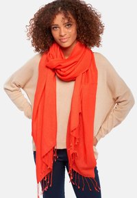 Écharpe rouge en tissu doux, drapée autour du cou avec des bords à franges, portée sur un haut beige. L'ensemble dégage une ambiance chaleureuse et confortable.