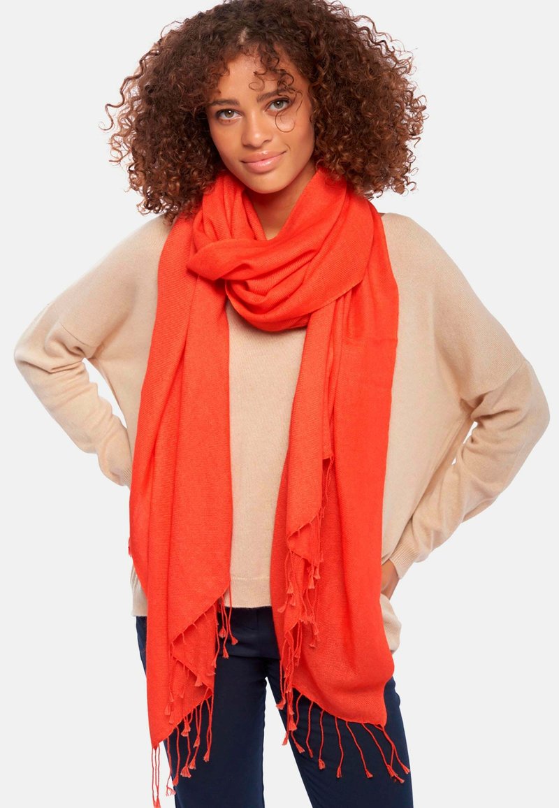 Écharpe rouge en tissu doux, drapée autour du cou avec des bords à franges, portée sur un haut beige. L'ensemble dégage une ambiance chaleureuse et confortable.