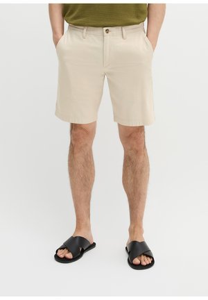 Mężczyzna ubrany w beżowe szorty, zielony teksturowany top i czarne sandały z paskami na krzyż, stoi z rękami w kieszeniach na białym tle.