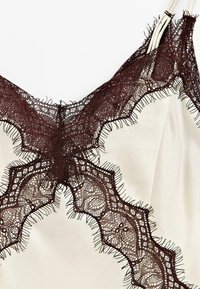 Satin-Camisole in Creme mit aufwendiger, dunkelbrauner Spitzenbordüre mit gewellten Kanten und fransigem Detail am Ausschnitt.