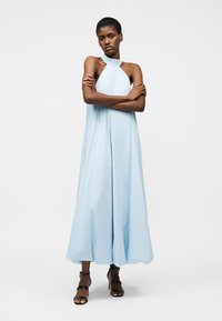 Femme debout, bras croisés, portant une robe longue sans manches bleu clair à col halter et des talons noirs à brides.