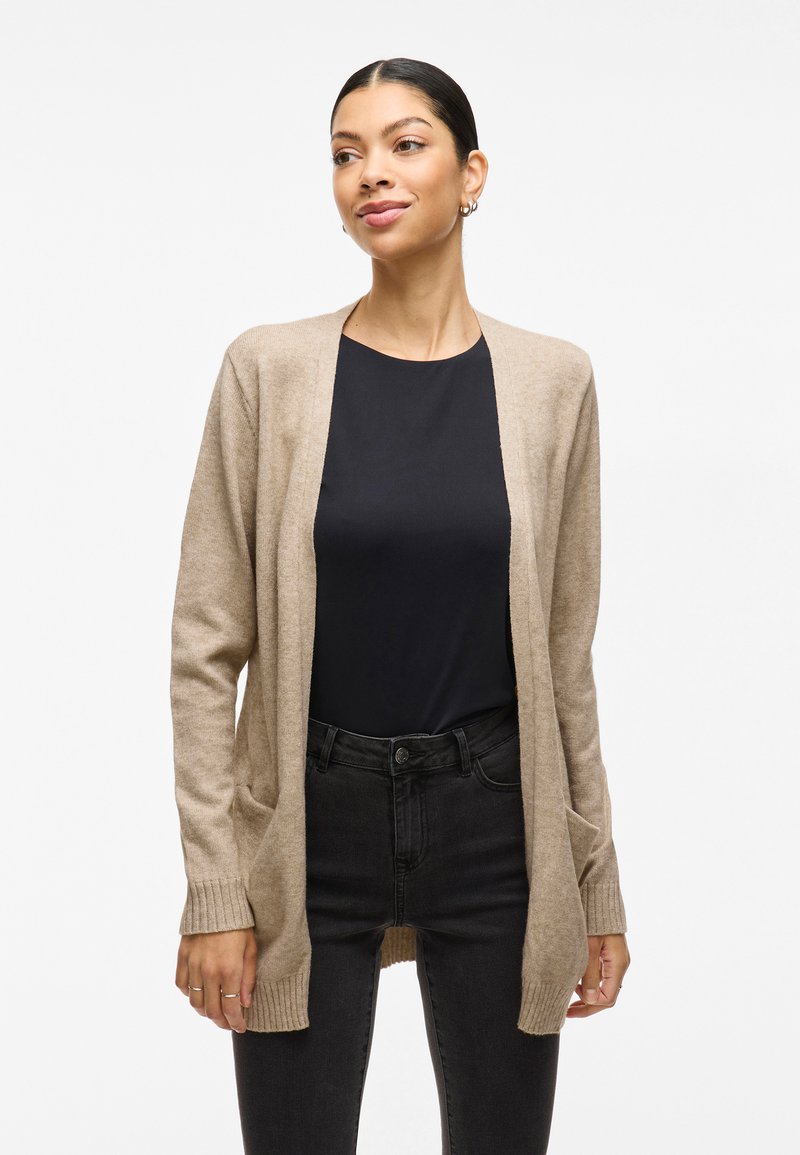 Beige cardigan med åpen front, ribbede mansjetter, og to frontlommer, laget av mykt strikket stoff. Brukt over en svart topp og mørke jeans.