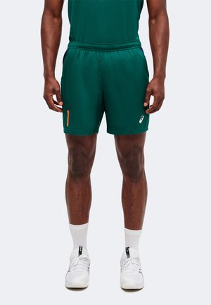 Man draagt groene sportshorts, witte sportschoenen en witte sokken tot halverwege de kuit, staand met ontspannen handen langs het lichaam.