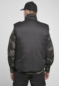 Svart ermeløs vest med høy krage og knappdetaljer, parret med en camo langermet skjorte. Stoffet virker glatt og holdbart.