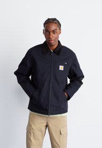 Veste bleu marine foncé avec fermeture éclair, col noir et poche poitrine arborant un logo. Portée par-dessus une chemise verte claire et un pantalon cargo beige.