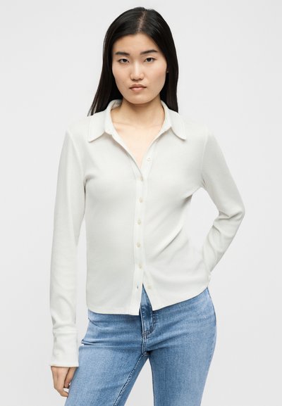 Νεαρή γυναίκα με μακριά σκούρα μαλλιά, φορώντας λευκό πουκάμισο με κουμπιά και μπλε τζιν, όρθια με ένα χέρι στη μέση.