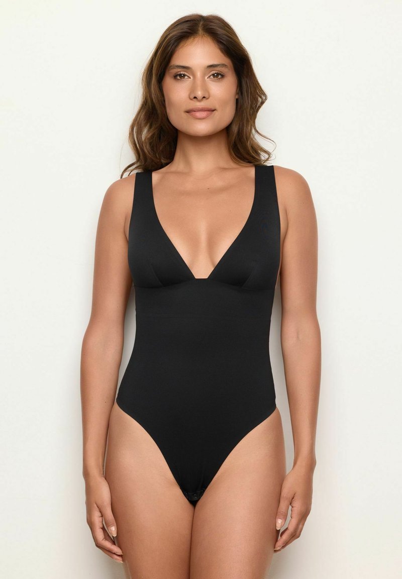 Kvinna med vågigt brunt hår klädd i en svart ärmlös bodysuit med djup V-ringning, stående mot en enfärgad ljus bakgrund.