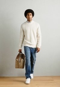 Suéter de punto color crema claro, jeans azules y zapatillas blancas. Lleva una bolsa de deporte de dos tonos en color marrón claro con acentos marrones y una superficie texturizada.