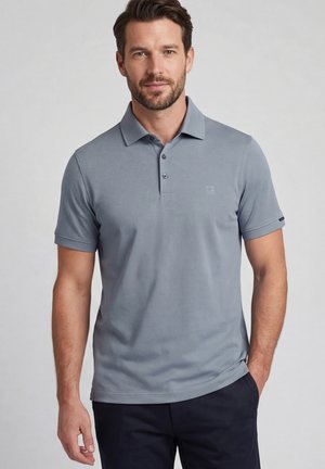 Homme debout, une main dans la poche, portant un polo bleu clair à manches courtes et un pantalon foncé sur un fond uni.