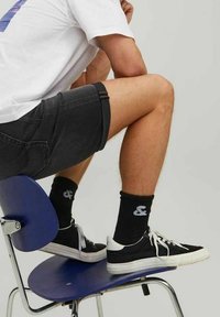 Calcetines negros con logo blanco, pantalones cortos negros y zapatillas negras con suela blanca, sentado en una silla azul, mostrando un atuendo casual.