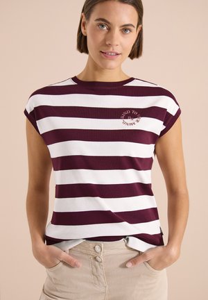Mujer con cabello corto y castaño, vistiendo una camiseta sin mangas a rayas granate y blanca y pantalones beige con las manos en los bolsillos.