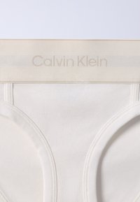 Λευκά εσώρουχα Calvin Klein με ριμπ ζώνη μέσης που διαθέτει διακριτικό μπεζ λογότυπο σε απλό φωτεινό φόντο.