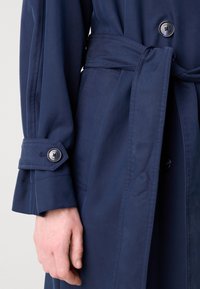 Trench-coat bleu navy avec une taille ceinturée, boutons double boutonnage et détails de poignets, fabriqué à partir d'un tissu lisse et texturé.