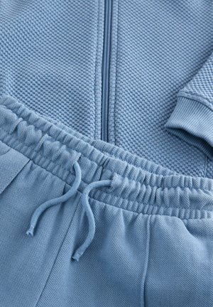 Ensemble de survêtement léger bleu clair en deux pièces comprenant une veste à zip texturée et un pantalon avec cordon de serrage à la taille. Le tissu est doux et possède des poignets et une taille côtelés.