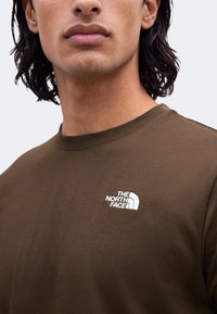 Homme aux cheveux mi-longs foncés porte un T-shirt vert olive avec le logo blanc "The North Face" sur la poitrine.
