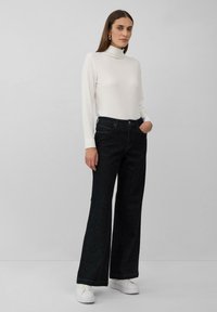 Pull à col roulé blanc côtelé associé à un jean flare en denim noir. Le mannequin porte des baskets blanches. Fond simple, axé sur les détails des vêtements.
