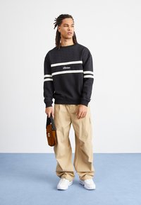 Felpa nera con strisce bianche e logo, pantaloni cargo beige, tiene una borsa marrone e indossa sneakers bianche. Sfondo semplice con pavimento blu.