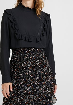 Zwarte blouse met ruches en knoopsluiting bij de hals, gecombineerd met een vloeiende zwarte bloemenrok met kleurrijke patronen.