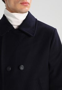 Peacoat di misto lana blu navy con ampi risvolti, chiusura doppiopetto e bottoni neri rotondi. Presenta un alto colletto sopra un dolcevita bianco.