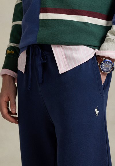 Tamsiai mėlyni sportinės kelnės su raišteliu liemens srityje, balti logotipai; derinami su žalia dryžuota džemperiu ir šviesiai rožine dryžuota marškinėlių apykakle.