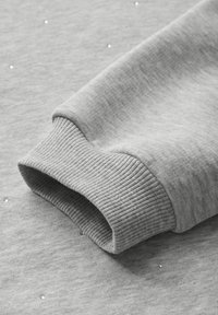 Detalle de sudadera gris que muestra un puño acanalado y pequeñas tachuelas decorativas en la manga, confeccionada en un tejido suave con una textura lisa.