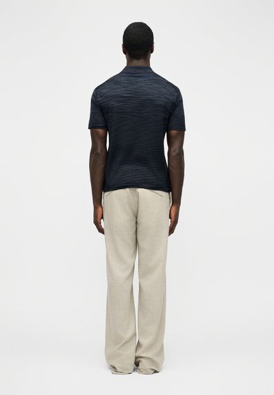 Άνδρας, φανερός από πίσω, που φοράει μπλε σκούρο πουκάμισο με κοντά μανίκια και χαλαρά ανοιχτό μπεζ παντελόνια, στέκεται μπροστά σε απλό λευκό φόντο.