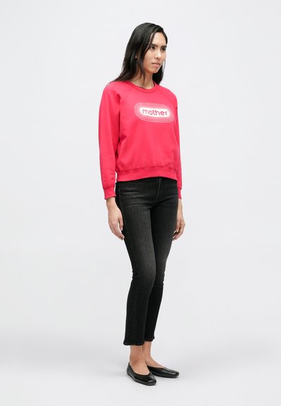 Raudonas džemperis su balta „mother“ logotipu, derinamas su tamsiomis siaurėjančiomis džinsais ir juodais bateliais. Medvilnės medžiaga, kasdienis dizainas, ribinės rankogaliai.