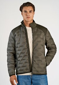 Olivgrön vadderad jacka med dragkedja, texturerade axeldetaljer och elastiska ärmmuddar, lager-på-lager över en krämfärgad stickad tröja.