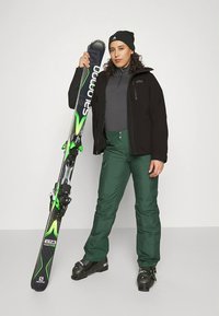 Ski equipment setup featuring a person in a black jacket, green ski pants, gray top, black beanie, holding green-accented skis with black bindings.