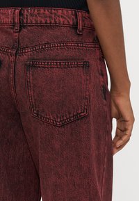 I pantaloni di jeans rossi presentano una finitura testurizzata, cuciture nere in contrasto e una tasca posteriore con un design a patta rettangolare.