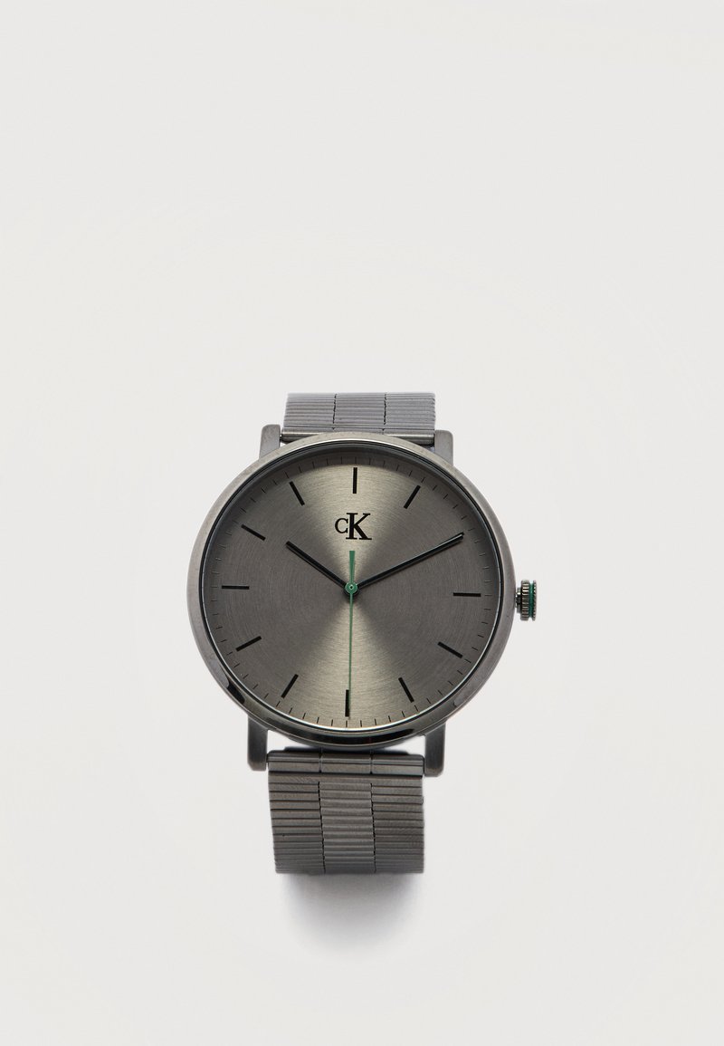 Montre Calvin Klein minimaliste en argent avec bracelet en maillons métalliques, index noirs et trotteuse verte sur fond neutre.