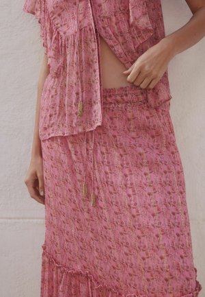 Woman wearing a pink floral patterned two-piece outfit with tassels, holding the fabric near her waist against a plain wall background.