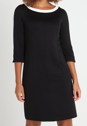 Kobieta ma na sobie czarną sukienkę do kolan z rękawami do łokcia, białym dekoltem oraz białymi paskami na ramionach i rękawach.