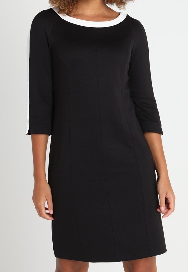 Femme portant une robe noire arrivant au genou, avec des manches trois-quarts, un col blanc et des rayures blanches le long des épaules et des manches.