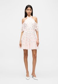 Donna che indossa un vestito rosa chiaro con scollo a barca, ricamo floreale e maniche corte trasparenti, abbinato a décolleté bianche a punta.