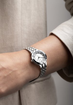 Montre-bracelet en métal argenté avec un cadran rond blanc, des chiffres romains et un bracelet argenté à motif. Associée à une manche en tissu beige.