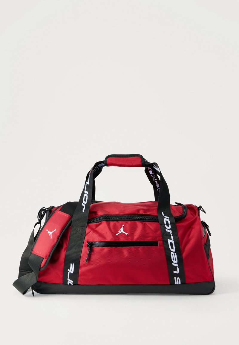 Sac de sport rouge avec des accents noirs, fabriqué en tissu durable. Comprend une poche zippée à l'avant, des poignées rembourrées et un marquage "JORDAN" audacieux sur les sangles.